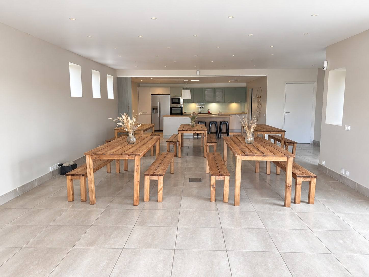 Salle de réception du Hameau des Sources, tables et bancs en bois dressés devant la cuisine équipée.