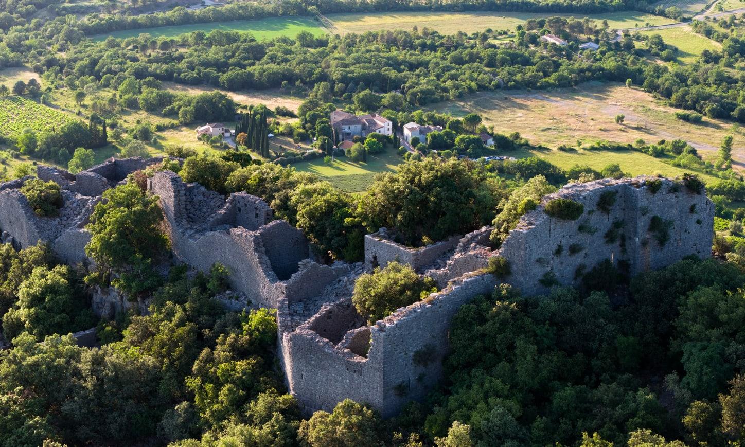 Vue aérienne d’un château en ruine et des mas en contrebas, au milieu de la garrigue.